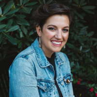 Adrienne Keene wearing a denim jacket in front of a verdant background. Headshot by Brittany Taylor.